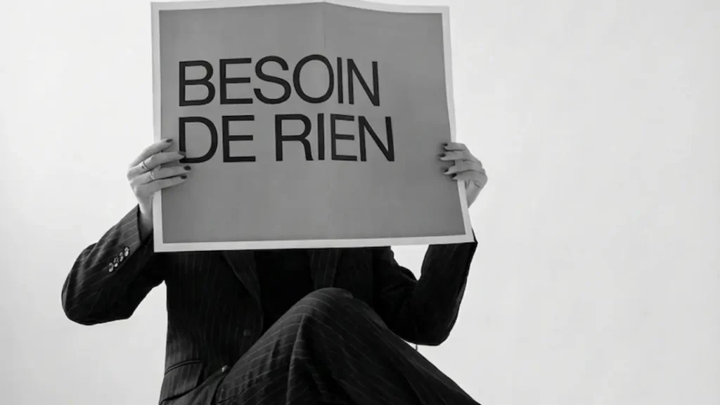 Femme assise habillée d'un costume noir tenant une feuille devant son visage avec pour note "besoin de rien"