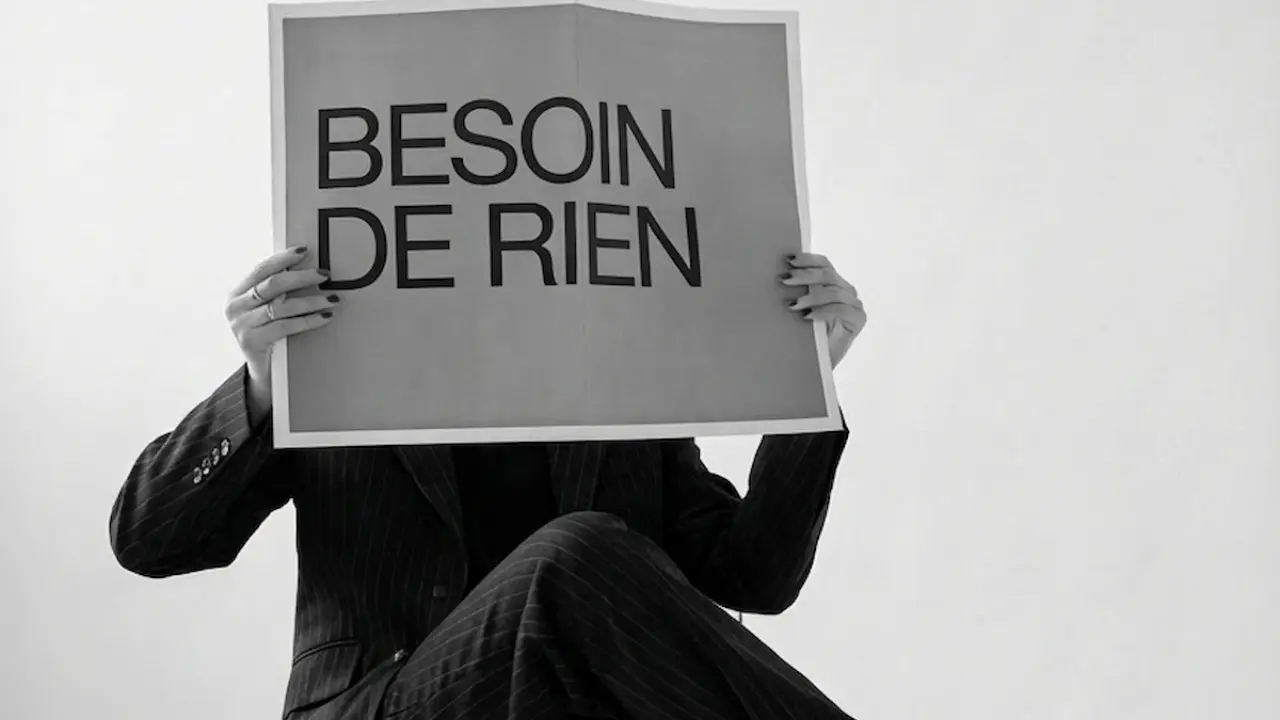 Femme assise habillée d'un costume noir tenant une feuille devant son visage avec pour note "besoin de rien"